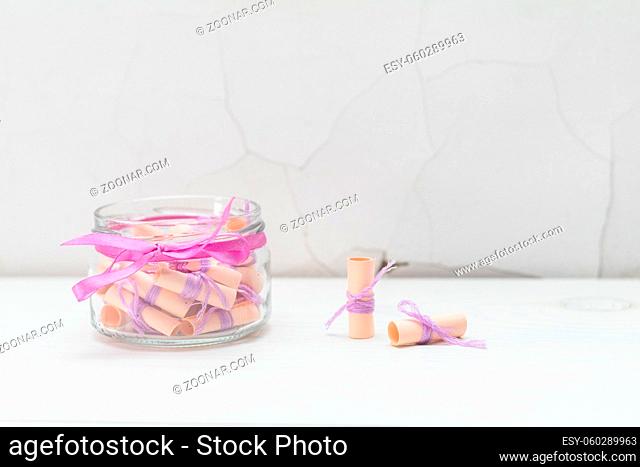 Paper wish scrolls witj violet threads in a glass jar with violet ribbon. White wooden surface close to the white cracked wall