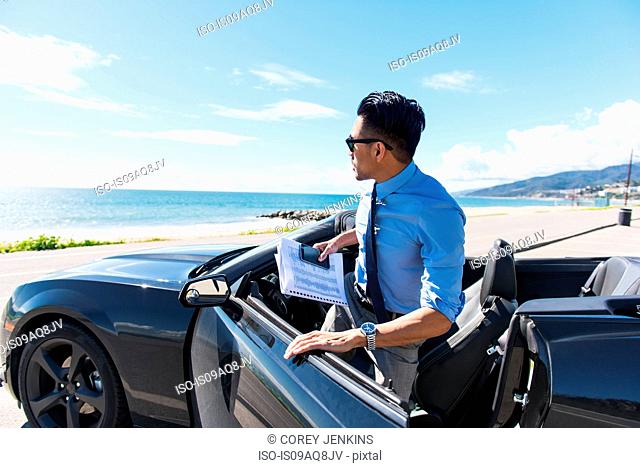 Young businessman getting out of car at coastal parking lot with paperwork