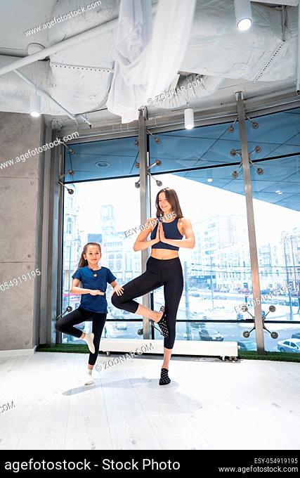 Young adult mother and little daughter together practicing yoga in the gym