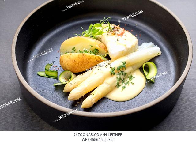 Modern German fried cod fish filet with white asparagus in hollandaise sauce und roast potatoes as top view on a plate
