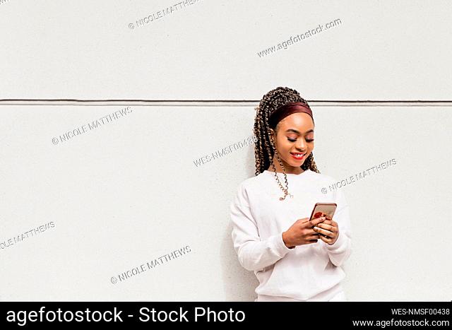 Smiling young woman using smart phone standing in front of white wall
