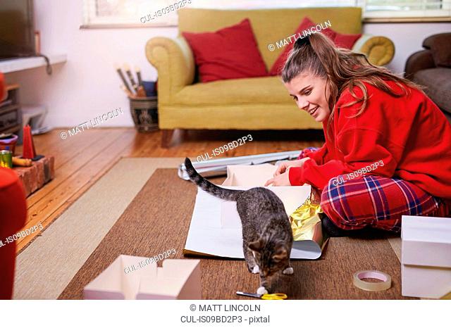 Young woman sitting on living floor wrapping gifts and watching cat