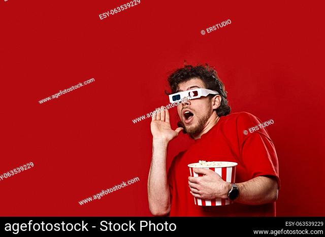 scared handsome bearded man in 3d-glasses holding bucket of popcorn on red background