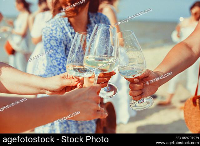 Happy female friends in summer dresses smiling and clinking glasses of wine while resting on beach together