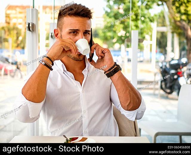 Close up of Handsome Young Man Drinking Coffee at the Shop While Talking on Cell Phone