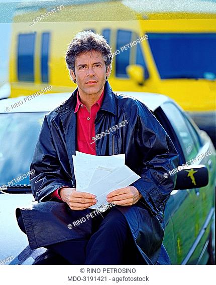 Italian song writer Claudio Baglioni (Claudio Enrico Paolo Baglioni) sittimg on a car's hood holding some letters. On the background there is the yellow van...