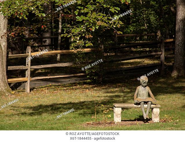 stone sculpture of a young girl sitting on a bench on a lawn with tall trees and a wood rail fence