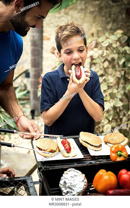 A boy and a man standing at an open barbecue eating