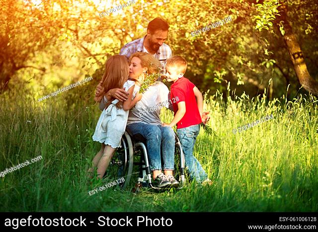 Cheerful Woman In Wheelchair Kisses Her Pretty Little Daughter And Hugs Son While Caring Man Looks Happily Over Them