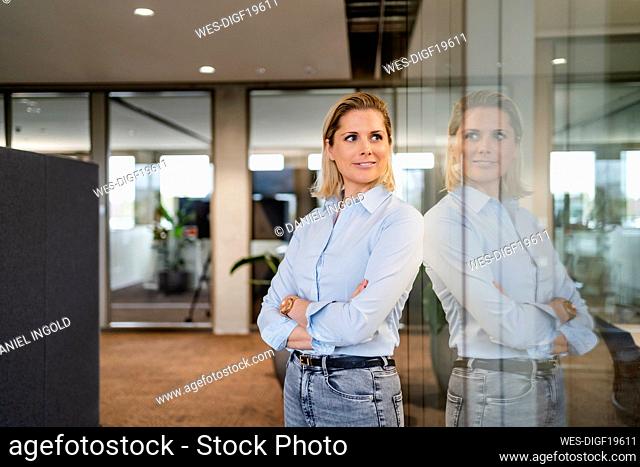 Thoughtful businesswoman with arms crossed standing by glass wall in office