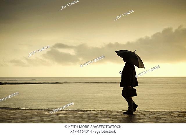 A man sheltering under an umbrella on a wet summer evening, Aberystwyth Wales UK
