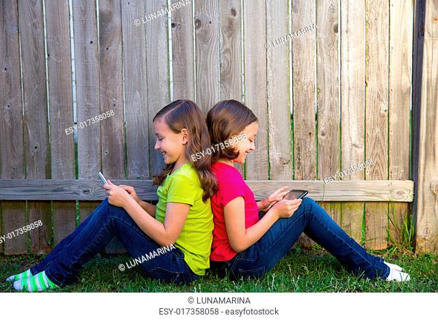 Twin sister girls playing with tablet pc sitting on backyard lawn fence leaning on her back