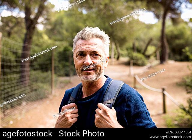 Smiling mature man with backpack standing in front of trees