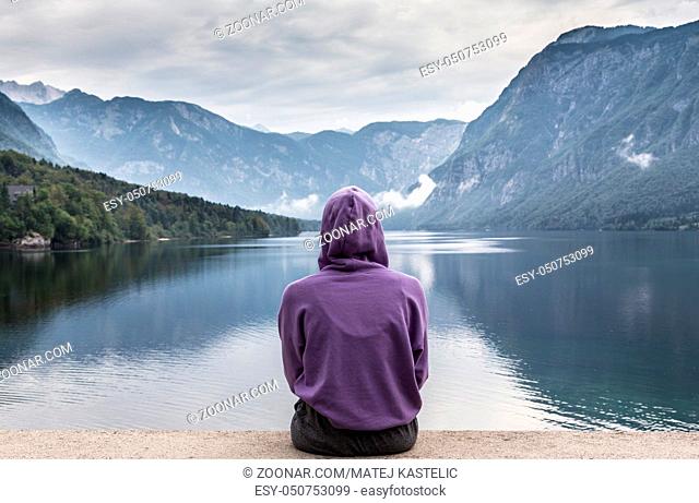 Solitary woman wearing purple hoodie watching tranquil overcast morning scene at lake Bohinj, Alps mountains, Slovenia