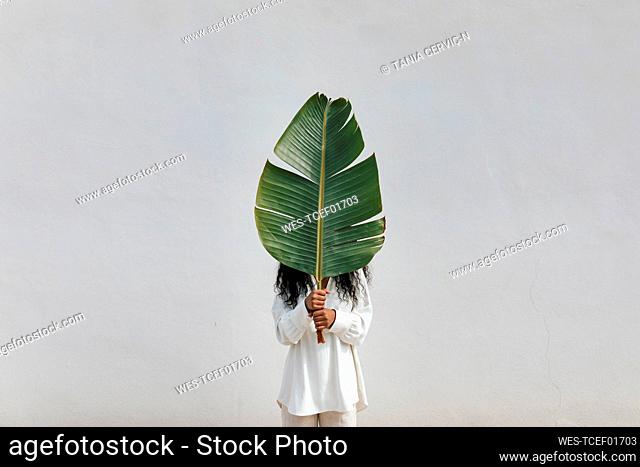 Woman covering face with big green leaf in front of white wall
