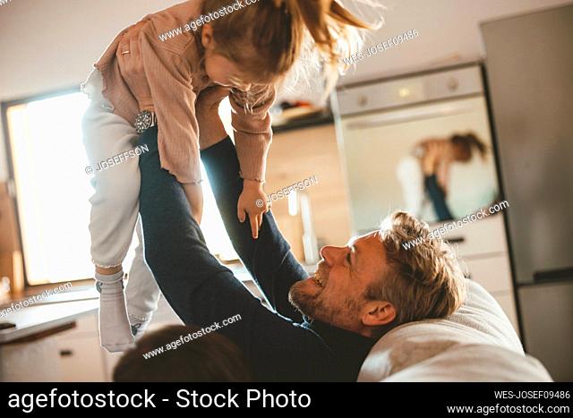 Happy man sitting on sofa holding daughter aloft at home