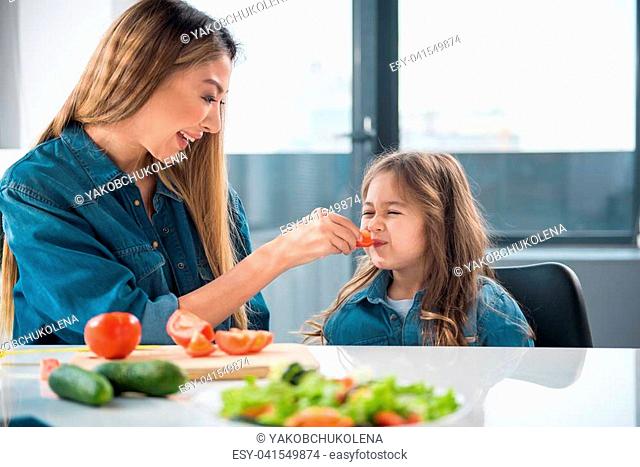 Smiling woman is giving piece of fresh vegetable to her daughter. The girl closing her mouth with aversion