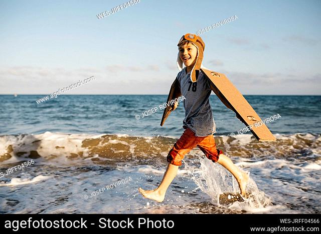 Boy with aircraft wings and cap splashing while running at beach