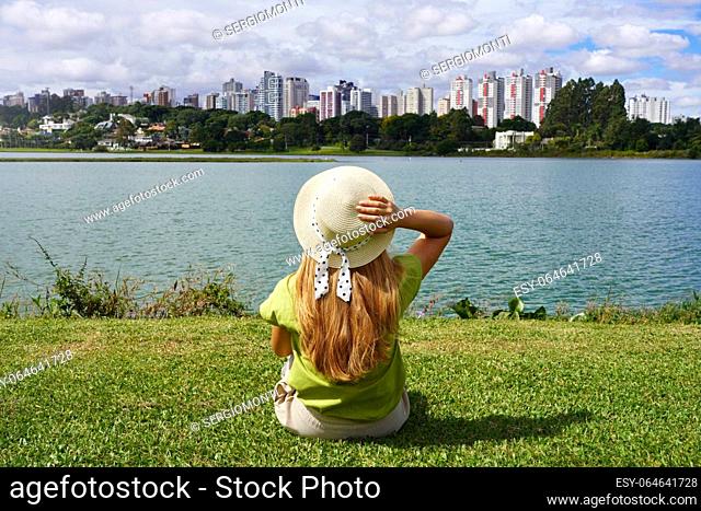 Girl with hat sitting on grass in Barigui Park enjoying Curitiba cityscape, Brazil