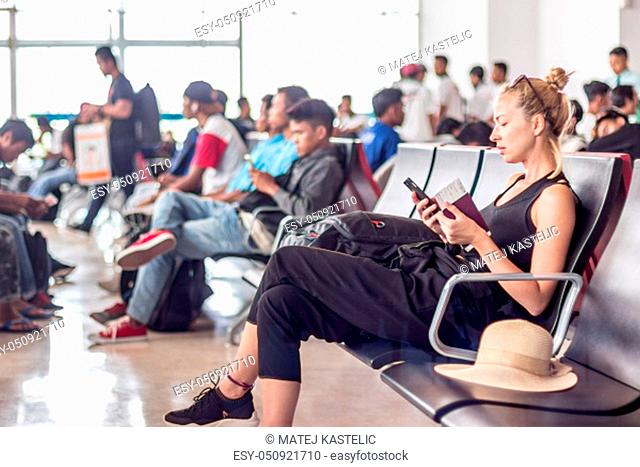 Casual sporty young blond female traveler using her cell phone while waiting to board a plane at the departure gates at the asian airport terminal