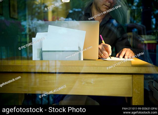 PRODUCTION - 01 October 2021, Bavaria, Munich: A man is sitting at a desk at home. He is writing with a pen on a piece of paper next to a laptop
