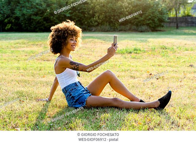 Tattooed young woman sitting on a meadow taking selfie with smartphone