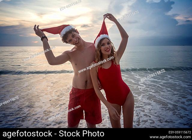 Christmas happy couple in red swimsuits smiling while standing on the beach over beautiful sunset background. Woman and man wearing santa hat enjoying travel...