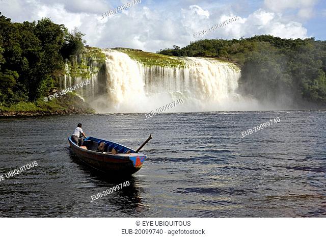 Canaima Village A Pemon tribe man while sailing with his boat at Canaima lake just in front of a big waterfall on a bright day with blue sky and white clouds
