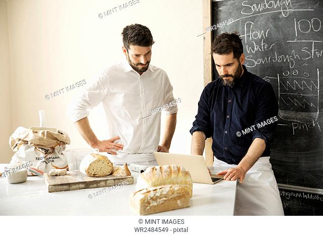 Two bakers standing at a table, using a laptop computer, freshly baked bread, a blackboard on the wall
