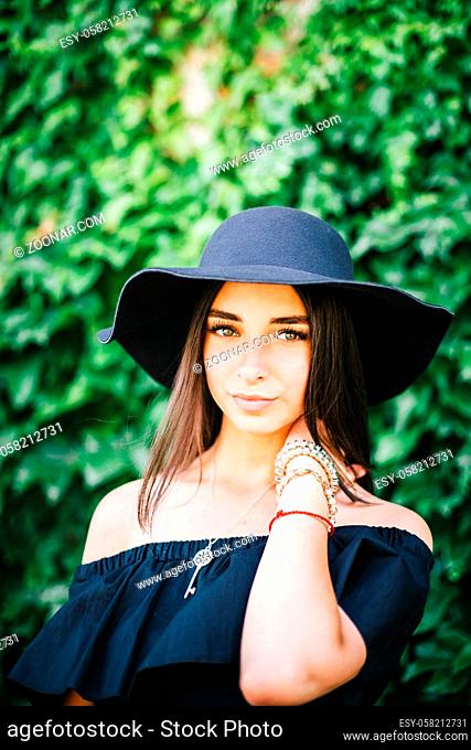 beautiful young sexy girl with swarthy skin and brunette with black hair dressed in a stylish black dress and hat on a background of green foliage