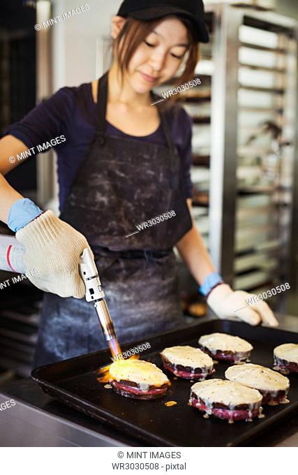 Woman working in a bakery, wearing oven gloves, using blowtorch, melting cheese on sandwiches