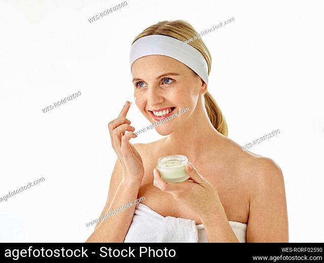 Young woman smiling while applying facial cream standing against white background