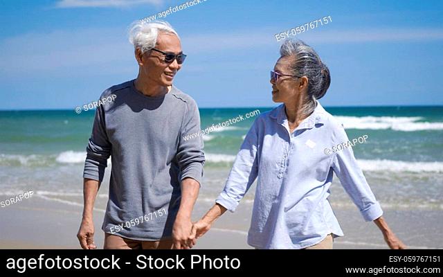 Happy Asian senior man and woman couple holding hands walking from the beach sunny with bright blue sky, Romantic elderly enjoy Travel summer vacation