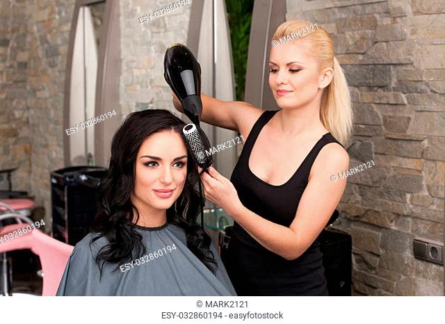 Beautician blow drying woman's hair after giving new haircut at parlor. blond girl drying dark-haired girl hair and smiling