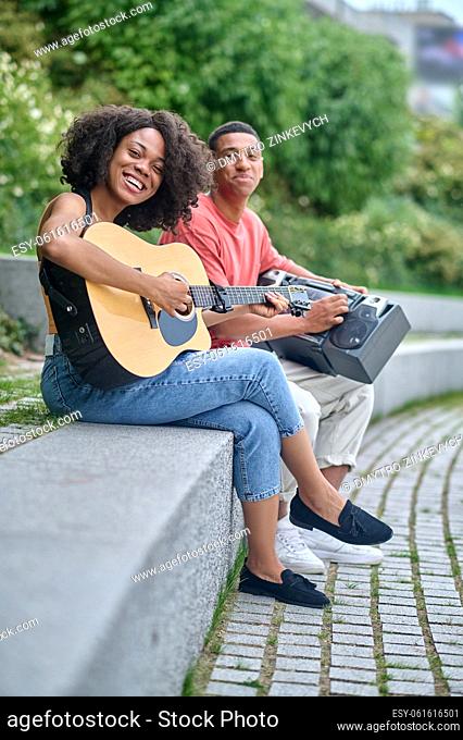 Good mood. African american girl playing guitar and guy with tape recorder sitting sideways smiling at camera in park