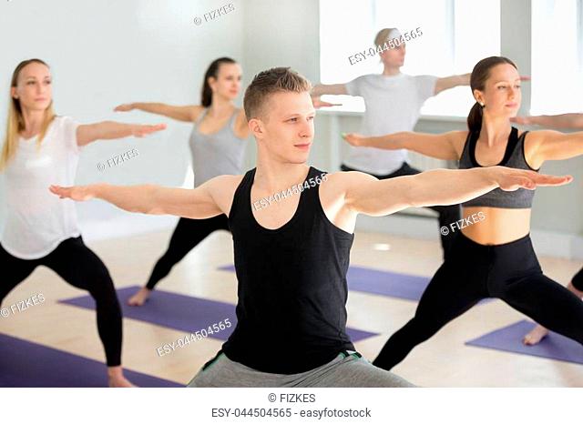 Group of young sporty people practicing yoga lesson, doing Warrior exercise, Virabhadrasana 2 pose, working out, indoor close up