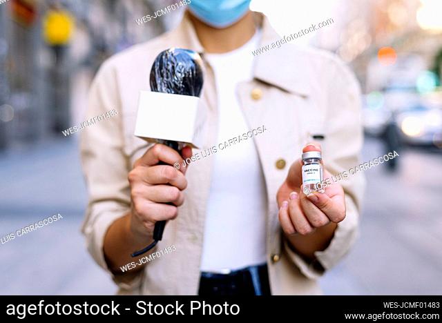 Close-up of female reporter wearing mask showing vaccine vial while standing outdoors