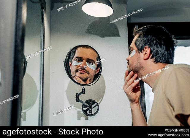 Tired man with under eye patches looking in mirror at bathroom