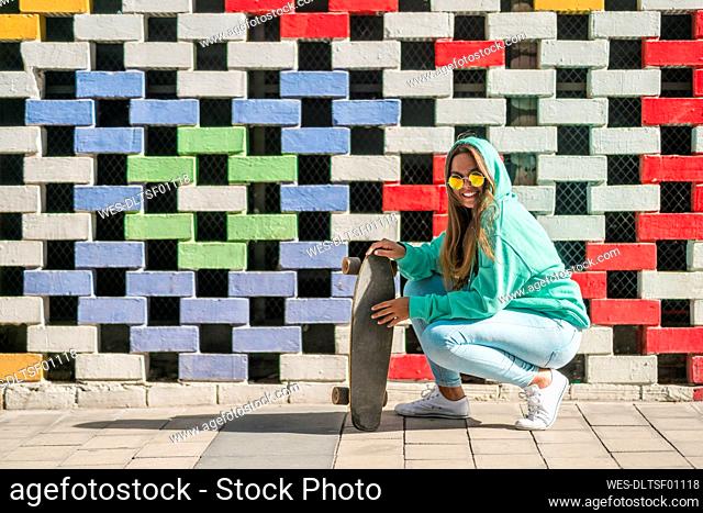 Young woman crouching while holding skateboard against colored wall