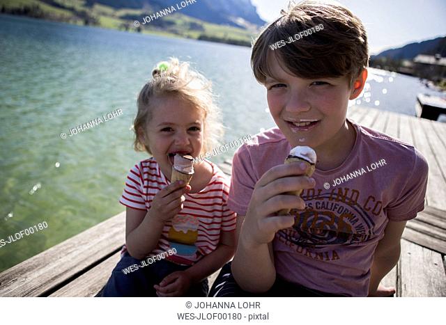 Austria, Tyrol, Walchsee, portrait of brother and sister sitting on jetty at a lake eating ice cream cones
