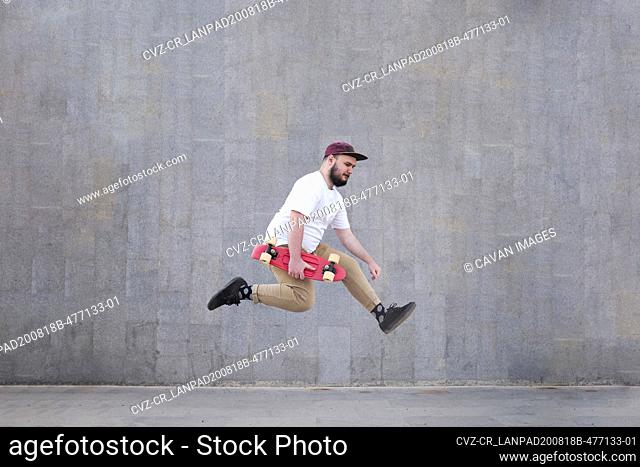 Young man jumping against a grey wall and holding his skateboard