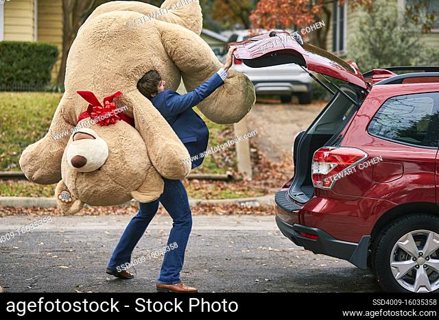 Wide shot of a man holding oversized teddy bear over his shoulder, closing the trunk of his car