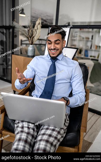 Home office. Gesturing attractive elegant man smiling working on laptop sitting in leather armchair at home