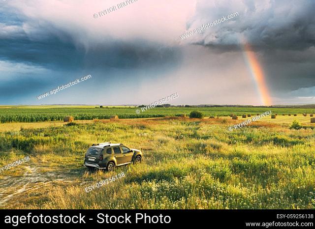 Elevated View Of Car SUV Parked Near Countryside Road In Summer Field Rural Landscape. Altered Rainy Sky With Rinbow Above Meadow Rural Landscape