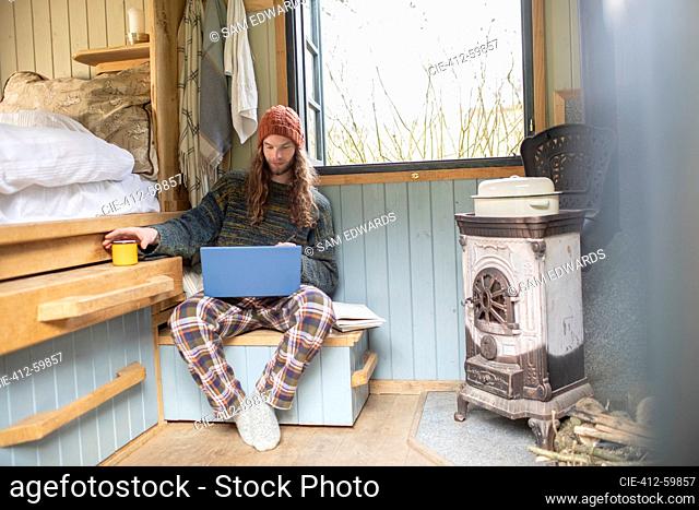 Young man in pajamas using laptop in tiny cabin rental
