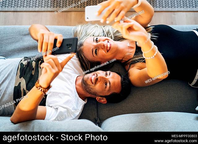 Portrait of laughing couple lying on the the couch taking selfies with smartphones