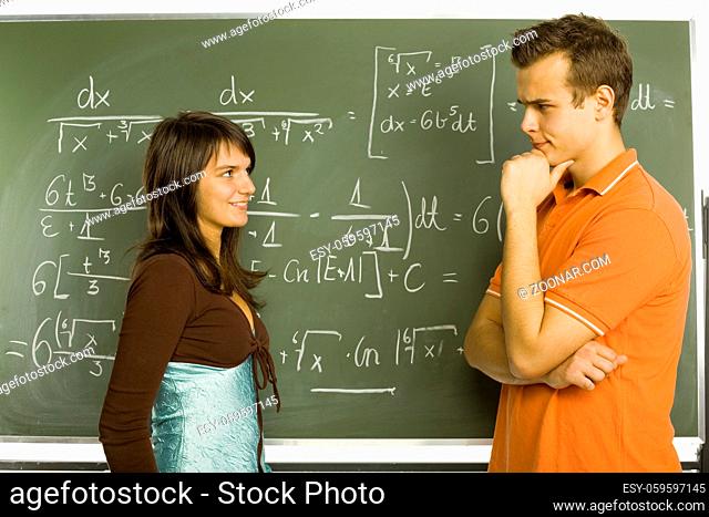 Two teenagers standing in front of blackboard. One smiling girl and thinking boy. Side view