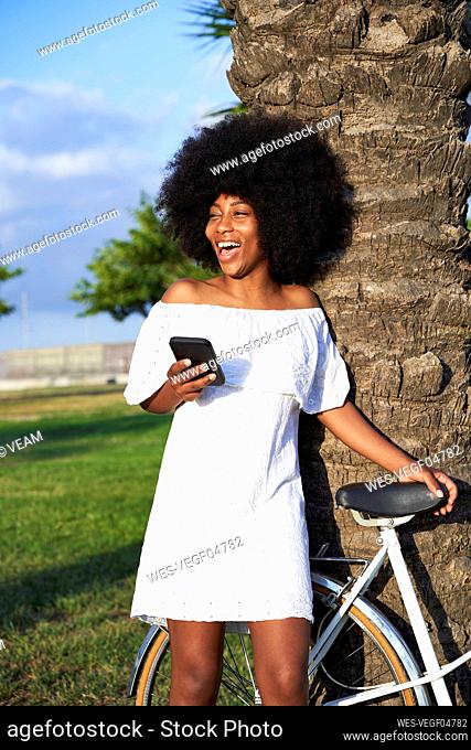 Afro woman laughing while standing with mobile phone by bicycle at park