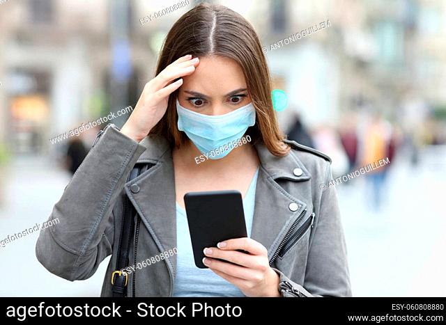 Front view portrait of worried woman with protective mask checking news on smart phone on a city street