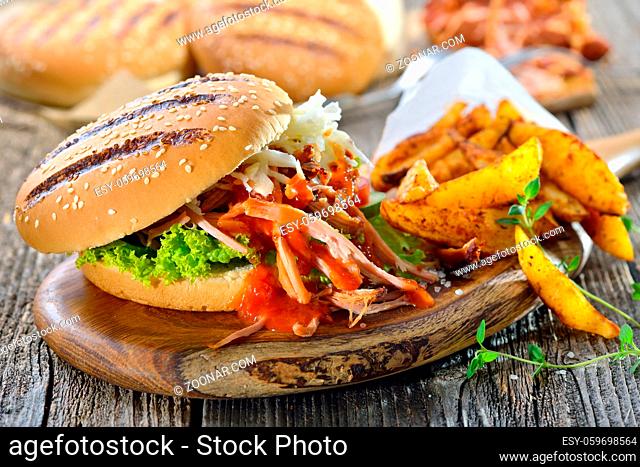 Barbecue pulled pork sandwich with coleslaw, hot BBQ sauce and potato wedges in a paper bag served on a wooden table